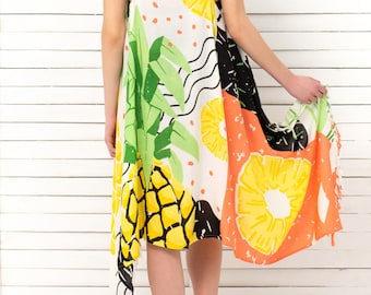 Könnte beinhalten: Ein ärmelloses, weißes Kleid mit einem lebendigen tropischen Fruchtdruck. Das Design zeigt große gelbe Ananasscheiben, grüne Blätter und orangefarbene Akzente. Das Kleid hat einen fließenden, asymmetrischen Saum und ist für warmes Wetter geeignet.