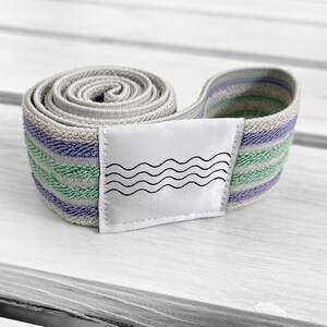 May include: A coiled fabric resistance band featuring horizontal stripes in shades of light purple, mint green, and beige. A white rectangular patch is stitched onto the center of the band, displaying a graphic print of four black wavy lines.