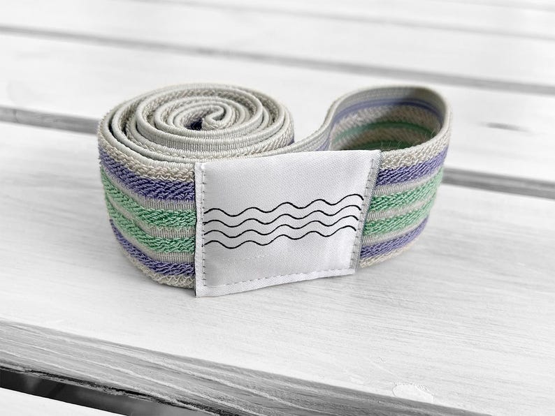 May include: A coiled fabric resistance band featuring horizontal stripes in shades of light purple, mint green, and beige. A white rectangular patch is stitched onto the center of the band, displaying a graphic print of four black wavy lines.
