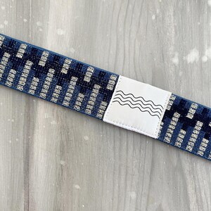 May include: A blue elastic strap featuring a repeating dark and light blue geometric pattern. A white rectangular patch is stitched onto the center of the strap, displaying a black line drawing of four horizontal wavy lines.