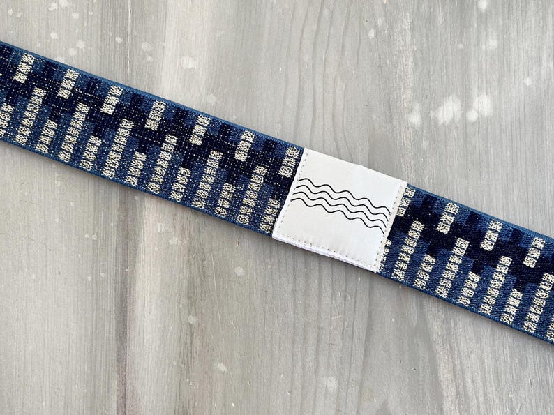 May include: A blue elastic strap featuring a repeating dark and light blue geometric pattern. A white rectangular patch is stitched onto the center of the strap, displaying a black line drawing of four horizontal wavy lines.