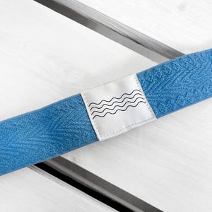 May include: A blue elastic headband featuring a textured, repeating leaf-like pattern across the fabric. A white rectangular fabric label is stitched onto the center, displaying a black graphic print of four horizontal, wavy lines.