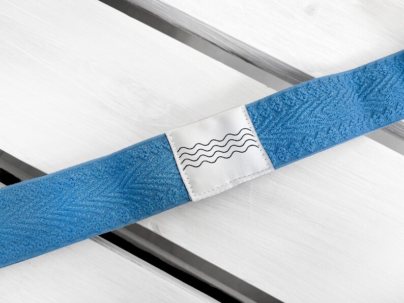 May include: A blue elastic headband featuring a textured, repeating leaf-like pattern across the fabric. A white rectangular fabric label is stitched onto the center, displaying a black graphic print of four horizontal, wavy lines.