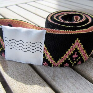 May include: A coiled black elastic strap featuring a repeating geometric diamond pattern in pink and yellow thread. A white rectangular fabric patch is attached to the strap, displaying a black graphic print of four horizontal wavy lines.