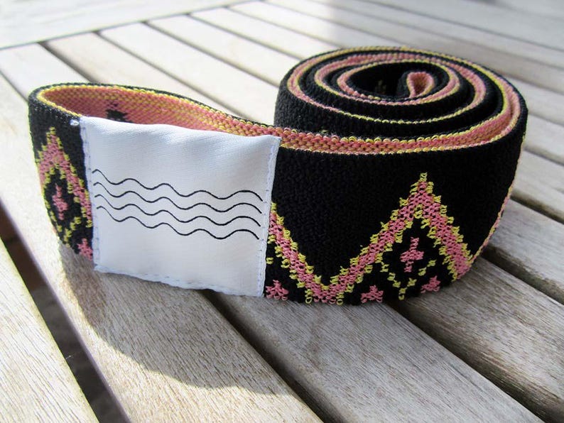 May include: A coiled black elastic strap featuring a repeating geometric diamond pattern in pink and yellow thread. A white rectangular fabric patch is attached to the strap, displaying a black graphic print of four horizontal wavy lines.