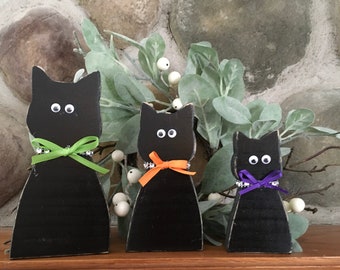 May include: Three black cat figurines with googly eyes and colorful ribbon collars. The cats are arranged in a row, each with a different ribbon color: green, orange, and purple. The background includes greenery and a stone wall.