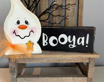May include: A wooden Halloween decoration featuring a smiling ghost with an orange nose and cheeks, a star, and an orange bow. A black sign with the word "Booya!" in white lettering sits next to the ghost. The items are on a small wooden bench.