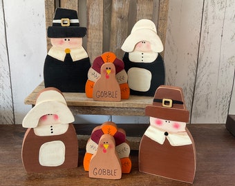 May include: Wooden Thanksgiving decorations featuring pilgrim figures and turkeys. The figures are painted in brown, black, and white, with details like hats and collars. The turkeys have colorful tail feathers and the word "GOBBLE" on their bodies. These decorative items are perfect for the fall season.