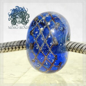 Könnte beinhalten: Eine blaue Glasperle mit einem silbernen und goldenen Gitterdesign und weißen Punkten. Die Perle befindet sich auf einem silbernen Armband.