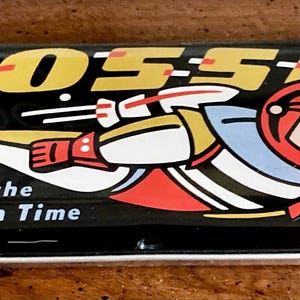 May include: A black metal tin with a white and red illustration of a rocket ship and astronaut. The text "FOSSIL" is in yellow letters and the text "WINNING the Race with Time" is in white letters.