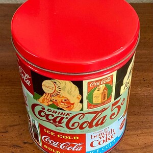 May include: A vintage Coca-Cola tin with a red lid. The cylindrical tin features various Coca-Cola advertisements, including the text "Drink Coca-Cola 5¢" and "Ice Cold." The design includes a child holding a Coca-Cola bottle.