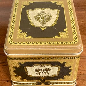 May include: A rectangular metal tin with a decorative design. The lid and sides are tan with gold trim and ornate scrollwork. The top features a dark brown panel with a gold crest. The bottom panel has a dark brown panel with the number 8.