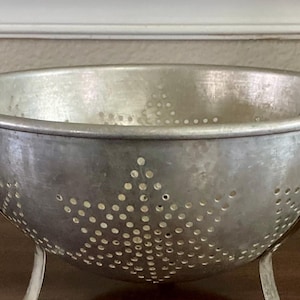 May include: A vintage, silver-colored metal colander with a wide, round bowl and small feet. The colander has numerous small holes and decorative star-shaped patterns of holes on the sides. It is designed for draining liquids from food items.
