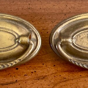 May include: Two oval-shaped brass drawer pulls with a decorative design. The pulls have a raised center section with a small hole in the middle.