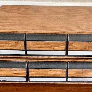 May include: A wooden storage box with six drawers. The drawers are black with a silver trim and have a wood grain pattern. The box is made of wood with a light brown stain.