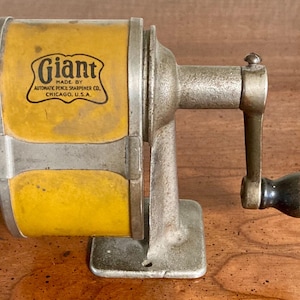 May include: Vintage pencil sharpener with a yellow and silver body. The sharpener has a crank handle with a black knob and the word "Giant" is printed on the front. Made by Automatic Pencil Sharpener Co., Chicago, U.S.A.