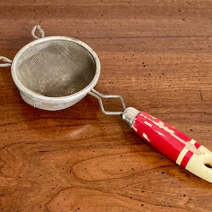 May include: A vintage metal strainer with a mesh screen and a red and white handle. The handle has a hole at the end for hanging. The strainer is likely used for sifting or straining ingredients in cooking or baking.