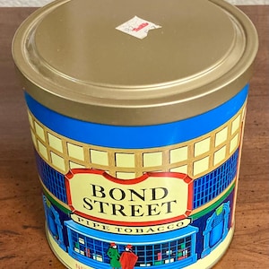 May include: A cylindrical tin of Bond Street Pipe Tobacco. The tin has a gold lid and a blue band. The main body of the tin features a yellow and blue illustration of a shop with the words "BOND STREET PIPE TOBACCO". Net weight is 12 oz.