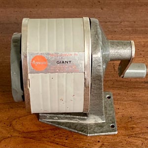 May include: A vintage manual pencil sharpener with a light gray cylindrical body and a textured dark gray metal base. The side features an orange circular logo with the text Apsco and the word GIANT printed in black capital letters on a silver plate.