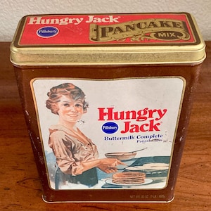 May include: Vintage Hungry Jack pancake mix tin. The brown tin features a red lid with gold trim and the words "Hungry Jack Pancake Mix." The front has an illustration of a woman making pancakes and the words "Hungry Jack Buttermilk Complete Pancake Mix."