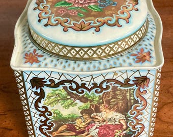 May include: A vintage square tin box with a floral design on the lid and a romantic scene on the side. The tin is decorated with gold trim and a blue and white chequered pattern.