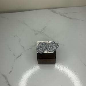 May include: A pair of silver-colored stud earrings shaped like the Millennium Falcon spaceship. The earrings are displayed on a small, dark wooden block with a white and gray marbled surface.
