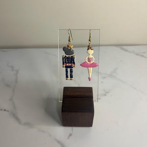 May include: A pair of dangle earrings featuring a nutcracker and a ballerina. The nutcracker is wearing a blue and gold uniform and the ballerina is wearing a pink tutu.