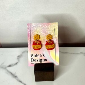 May include: A pair of red and yellow "Hunny" earrings. The earrings are shaped like honey pots with a honeycomb design on top. The earrings are displayed on a card with the text "Shlee's Designs".