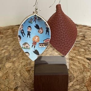 May include: A pair of dangle earrings with a blue and white patterned design featuring cartoon characters and a motel sign. The other earring is solid brown leather.