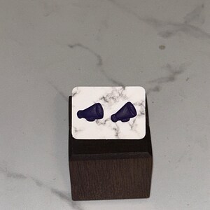 May include: A pair of small, blue megaphone earrings on a white marble display stand. The stand is on a dark brown wooden base.