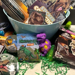 May include: A blue basket filled with dinosaur-themed toys and treats. The basket includes a purple dinosaur stress ball, a bag of dinosaur fruit snacks, a sticker book, a dinosaur puzzle, and a dinosaur-themed bubble wand.