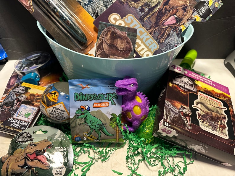 May include: A blue basket filled with dinosaur-themed toys and treats. The basket includes a purple dinosaur stress ball, a bag of dinosaur fruit snacks, a sticker book, a dinosaur puzzle, and a dinosaur-themed bubble wand.