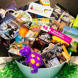 May include: A blue metal bucket filled with dinosaur-themed toys and gifts. The items include a purple dinosaur stress ball, a sticker and activity book, a dinosaur puzzle, a dinosaur-shaped cookie cutter, and a dinosaur-themed bath bomb.