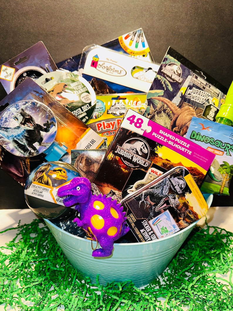 May include: A blue metal bucket filled with dinosaur-themed toys and gifts. The items include a purple dinosaur stress ball, a sticker and activity book, a dinosaur puzzle, a dinosaur-shaped cookie cutter, and a dinosaur-themed bath bomb.