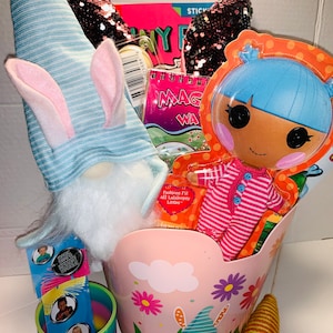 May include: A pink plastic basket with a gnome and flowers printed on it. The basket is filled with a variety of items, including a blue and white striped gnome hat, a Lalaloopsy doll, a book, a notepad, and a rainbow slinky.
