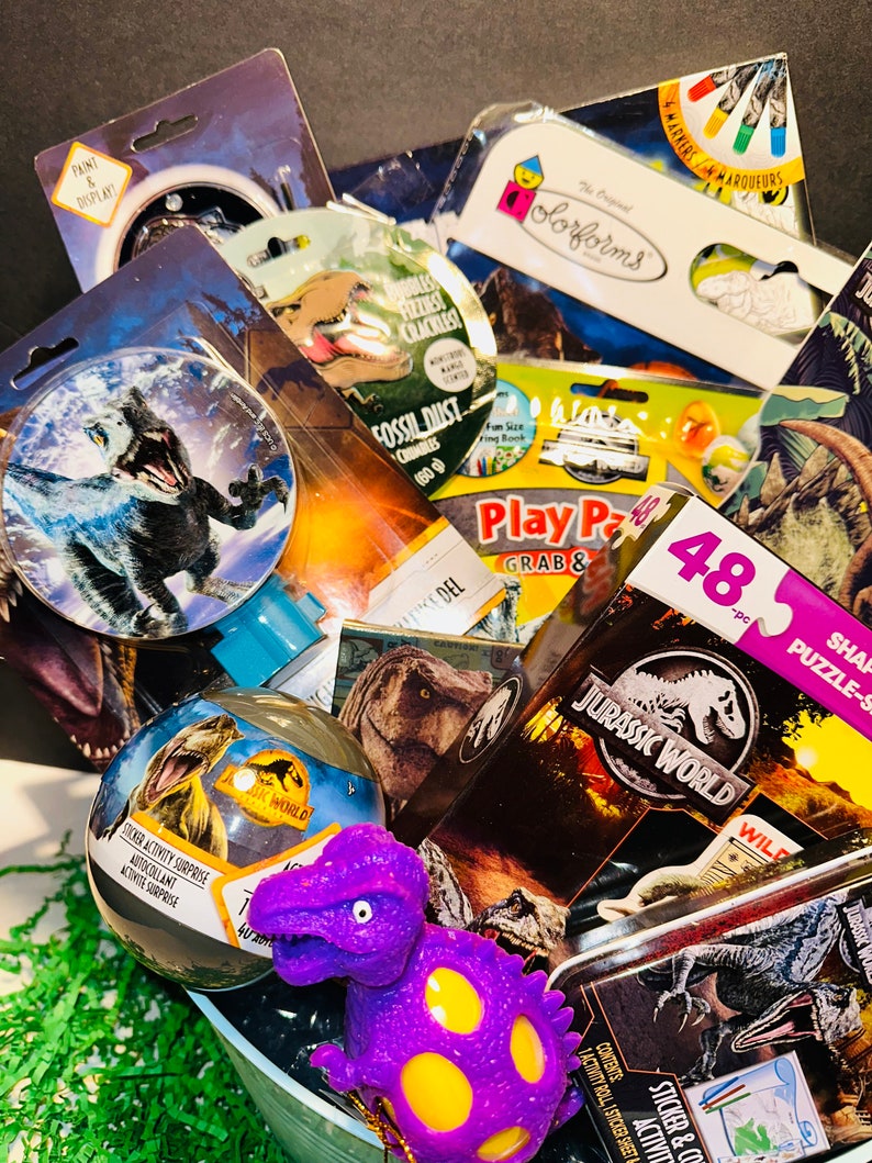 May include: A gift basket filled with dinosaur-themed toys and activities. The basket includes a purple dinosaur toy, a Jurassic World sticker activity, a dinosaur puzzle, and a dinosaur-shaped play dough set.