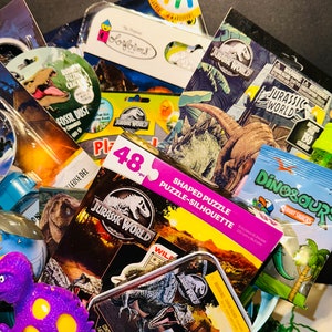 May include: A green basket filled with dinosaur-themed toys and activities. The basket includes a 48-piece shaped puzzle, a sticker and color activity book, a Jurassic World tin, a dinosaur-shaped fruit snack, and a purple dinosaur toy.