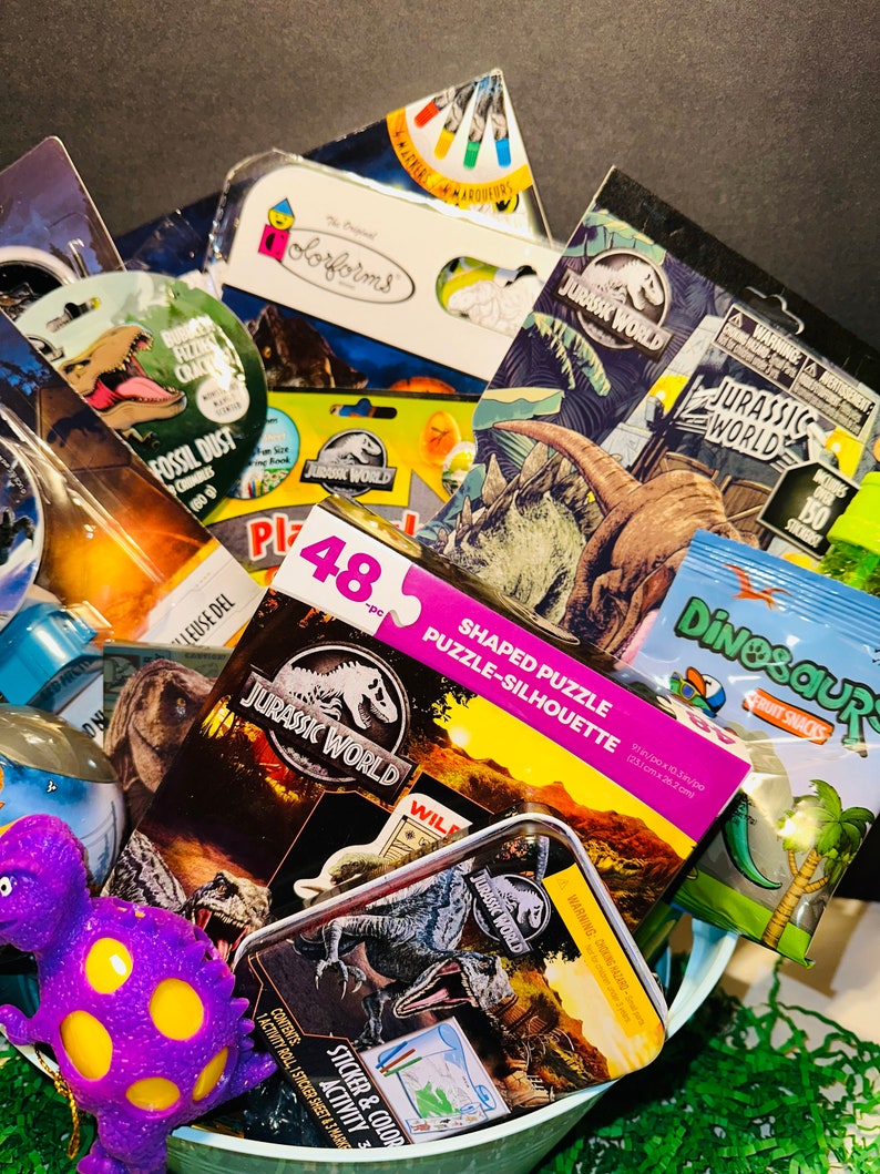 May include: A green basket filled with dinosaur-themed toys and activities. The basket includes a 48-piece shaped puzzle, a sticker and color activity book, a Jurassic World tin, a dinosaur-shaped fruit snack, and a purple dinosaur toy.