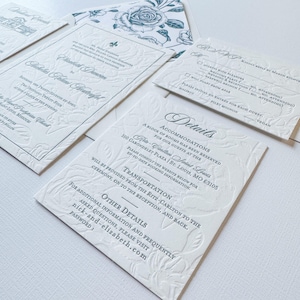 May include: Elegant white wedding invitation suite with embossed floral details and teal text. Includes invitation, RSVP card, details card, and envelope with a floral design. The invitation includes the names Nicholas and Elizabeth.