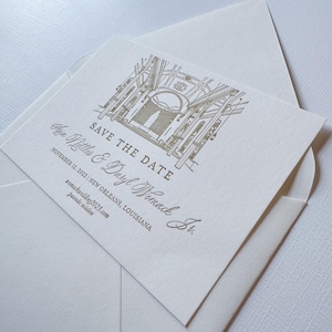 May include: A cream-colored Save the Date card with a gold architectural illustration, nestled inside a matching envelope. The card features the names Asia Nettles & Daryl Womack Jr., the date November 12, 2023, and New Orleans, Louisiana.