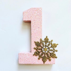 Pode incluir: N&uacute;mero um de glitter rosa com uma decora&ccedil;&atilde;o de floco de neve de glitter dourado.