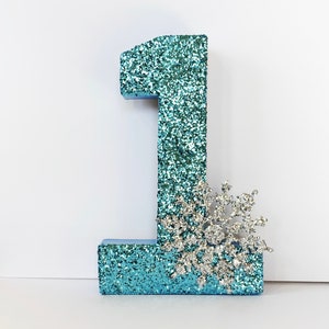 Pode incluir: Um n&uacute;mero um de glitter azul turquesa com uma decora&ccedil;&atilde;o de floco de neve de glitter prateado.