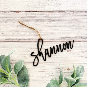 May include: Black wooden ornament with the name "Shannon" written in cursive script. The ornament is hanging from a twine string and is on a white wood background.
