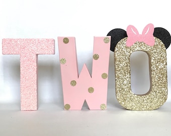 May include: Pink and gold glitter letters spelling out the word "TWO" with gold polka dots on the "W" and a pink bow and black ears on the "O".