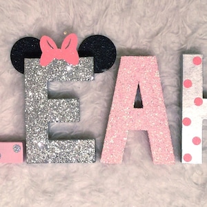 Op de afbeelding: Roze, zilveren en witte glitterletters die de naam "LEAH" spellen met zwarte en roze Minnie Mouse oren bovenop de "E".