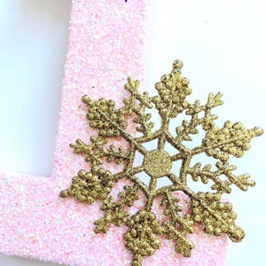 Pode incluir: Uma letra "1" de glitter rosa com um floco de neve de glitter dourado.