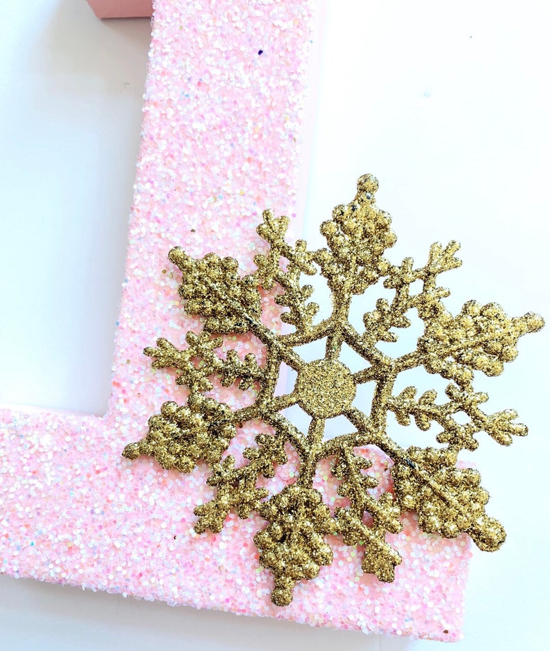 Pode incluir: Uma letra "1" de glitter rosa com um floco de neve de glitter dourado.