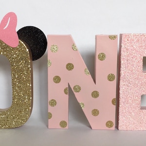 Op de afbeelding: Goudkleurige glitterletter "O" met een roze strik en zwarte oren, roze en gouden stippen op de letters "N" en "E", en een effen roze glitterletter "E" die samen het woord "ONE" vormen.
