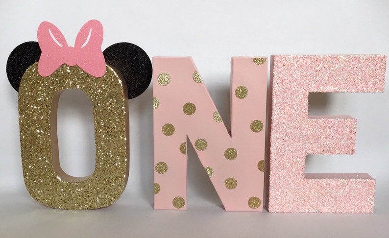 Op de afbeelding: Goudkleurige glitterletter "O" met een roze strik en zwarte oren, roze en gouden stippen op de letters "N" en "E", en een effen roze glitterletter "E" die samen het woord "ONE" vormen.