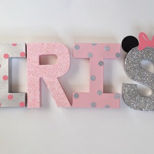 Op de afbeelding: Roze en zilveren glitterletters die de naam "IRIS" spellen met een zilveren glitterletter "S" met zwarte muizenoren en een roze strik.